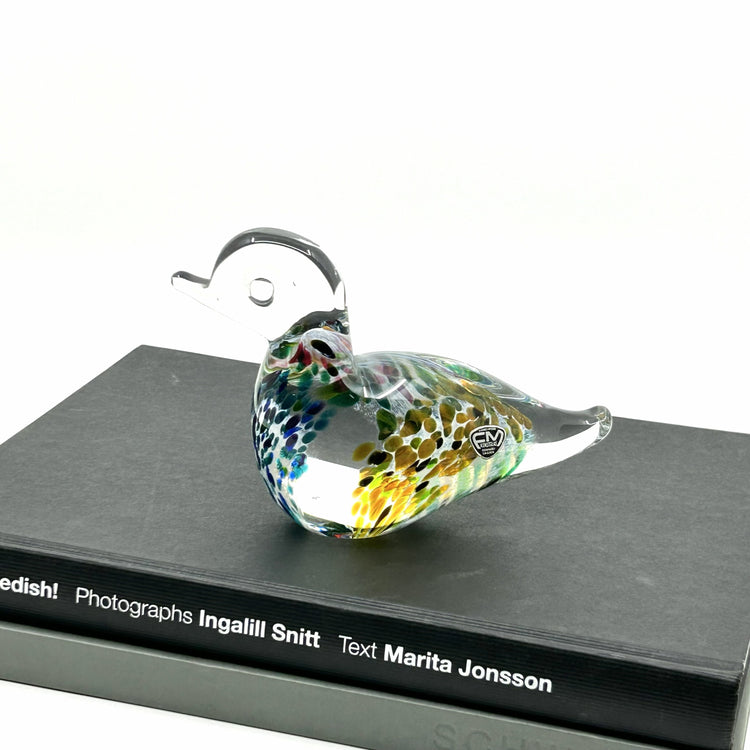 Josef Marcolin Vogel-Figur aus Glas: Handgefertigte Glasente mit farbenfrohen, konfettiartigen Einschlüssen im Glas auf schwarzem Buch – skandinavische Glaskunst aus Ronneby, Schweden