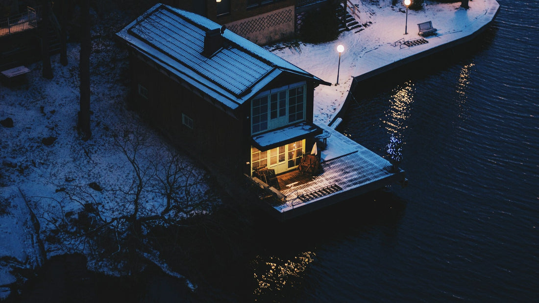 Nordic-Noir-Stimmung in Schweden: beleuchtetes Haus am Wasser im Winter, Schnee, Dunkelheit und reflektierendes Licht