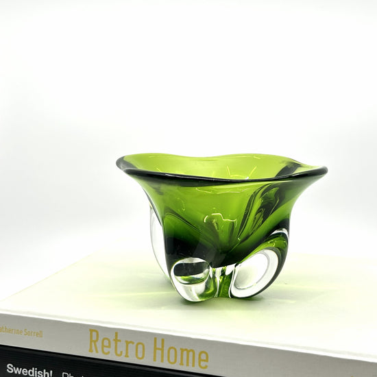 Magnor Dekoschale auf Buch 'Retro Home': Olivgrüne Glas-Schale mit organischen Mustern, platziert auf einem Buch über skandinavisches Retro-Design – norwegische Handwerkskunst